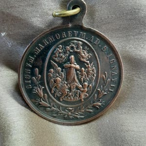 Könnte beinhalten: Runde, antike Bronzemedaille mit Aufhängeschlaufe. Die Medaille zeigt ein zentrales Relief einer bekleideten Figur, umgeben von Engeln, und die Inschrift "SIGNVM.MARMOREVM.AD.SIGNATI". Dekorative florale Elemente umrahmen das zentrale Bild.