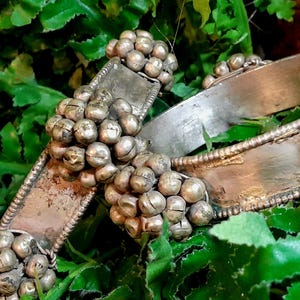 Può includere: Primo piano di un braccialetto argentato con grappoli di piccole perline rotonde. Il braccialetto ha un design rettangolare con dettagli intricati. Il gioiello è posto su uno sfondo di fogliame verde, creando un contrasto naturale.