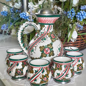 Könnte beinhalten: Ein Vintage-Teeservice aus Keramik mit einer großen Kanne mit Metalldeckel und sechs passenden Tassen. Das Set ist mit Blumenmustern in Rot, Grün und Braun auf weißem Grund verziert. Die Kanne hat einen geschwungenen Griff.