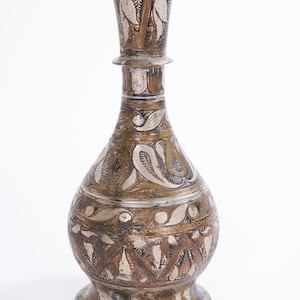 Puede incluir: Un jarrón de metal antiguo con intrincados diseños grabados. El jarrón presenta un cuerpo bulboso, un cuello delgado y un borde acampanado. El metal tiene una combinación de colores plateado y bronce, con patrones florales detallados.