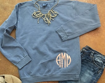 May include: A light blue sweatshirt with a gold necklace and a monogrammed "AMM" in a circle on the left side. The sweatshirt is paired with blue jeans and brown canvas shoes.