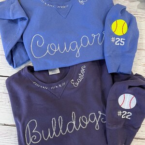 May include: Two crewneck sweatshirts, one periwinkle and one dark purple. The periwinkle sweatshirt has "Cougar" stitched across the front and "Reese" on the collar. The dark purple sweatshirt has "Bulldogs" stitched across the front and "Easton" on the collar. Both have sports-themed patches.