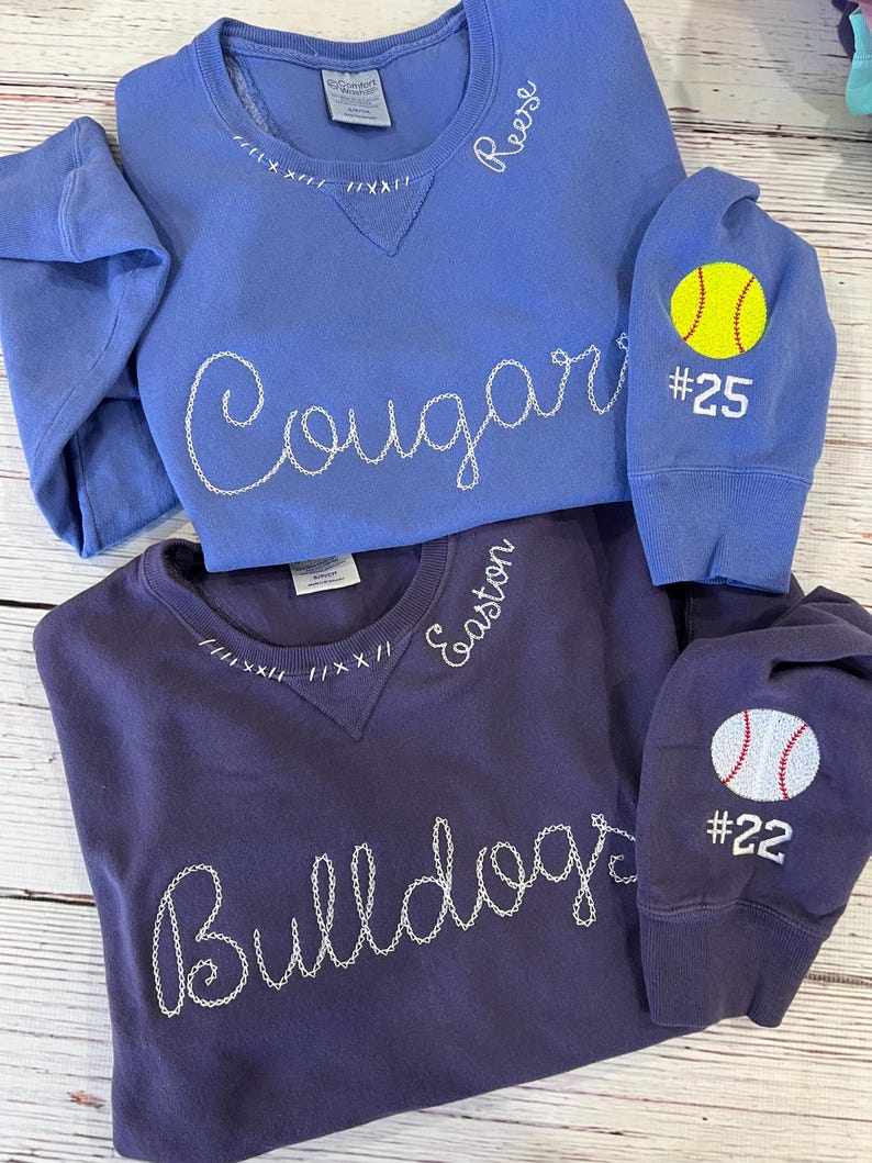 May include: Two crewneck sweatshirts, one periwinkle and one dark purple. The periwinkle sweatshirt has "Cougar" stitched across the front and "Reese" on the collar. The dark purple sweatshirt has "Bulldogs" stitched across the front and "Easton" on the collar. Both have sports-themed patches.