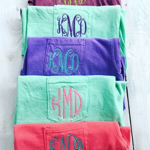 May include: Five t-shirts stacked on top of each other. Each t-shirt has a different color and a pocket on the back with the monogram "KMD" embroidered in a different color thread.