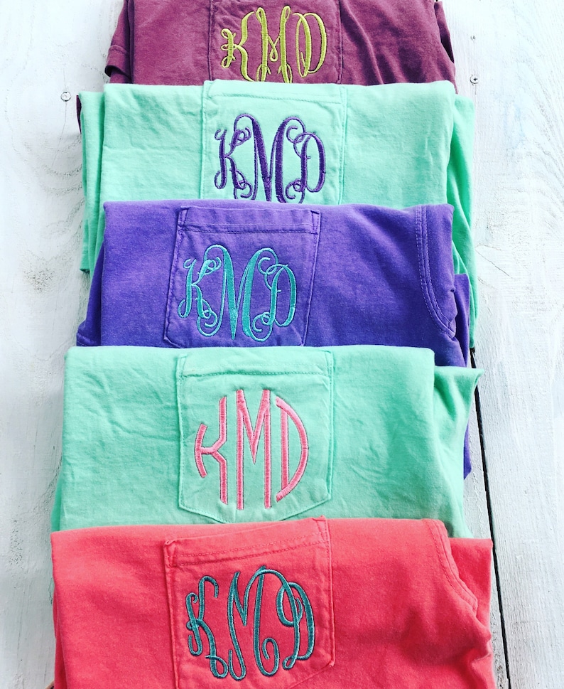 May include: Five t-shirts stacked on top of each other. Each t-shirt has a different color and a pocket on the back with the monogram "KMD" embroidered in a different color thread.