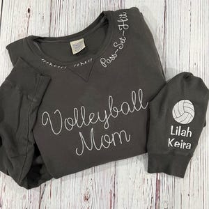 May include: A dark gray sweatshirt with the text "Volleyball Mom" and "Pass-Set-Hit" embroidered on the front. The left sleeve has a white volleyball and the text "Lilah Keira" embroidered on it.