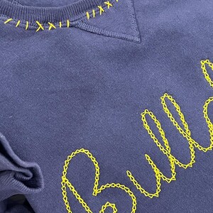 May include: A navy blue sweatshirt with yellow embroidered stitching. The neckline features decorative yellow stitching, and the word "Bull" is embroidered in a cursive font. The sweatshirt is a size S/P/CH.