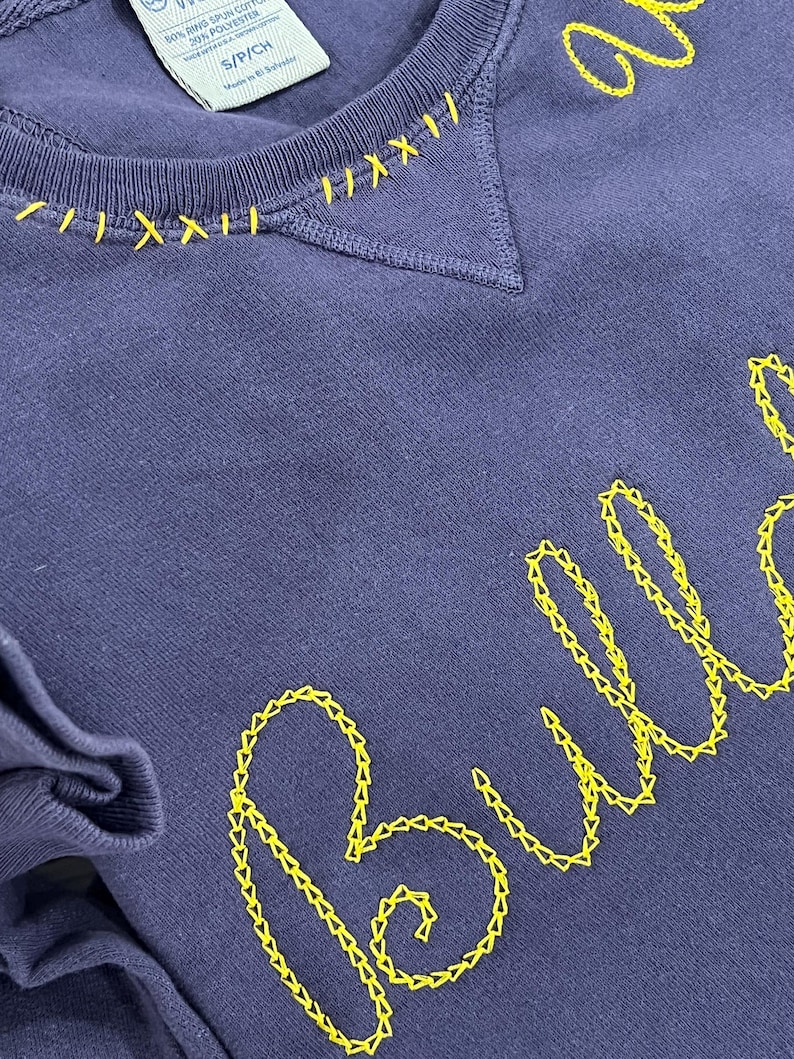 May include: A navy blue sweatshirt with yellow embroidered stitching. The neckline features decorative yellow stitching, and the word "Bull" is embroidered in a cursive font. The sweatshirt is a size S/P/CH.