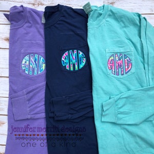 以下が含まれることがあります： 前面にポケットが付いた長袖Tシャツが3枚。ポケットにはカラフルなモノグラムのデザインが施されています。シャツの色はパープル、ネイビーブルー、ターコイズです。