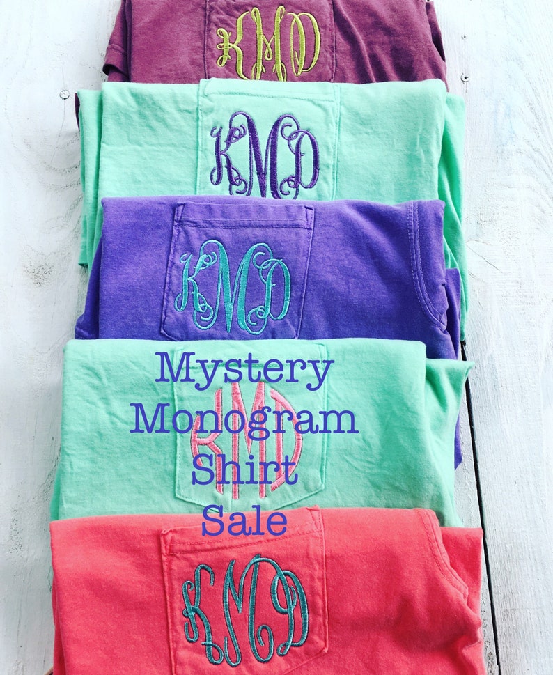 May include: Stack of five t-shirts with different colors and embroidered monograms in the pocket. The text "Mystery Monogram Shirt Sale" is printed on the shirts.