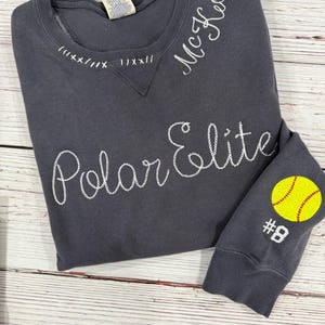 May include: A dark grey sweatshirt with white embroidered text reading "Polar Elite" and "McKeon." The sleeve features a yellow softball with red stitching and the number 8. The neckline has decorative white stitching.