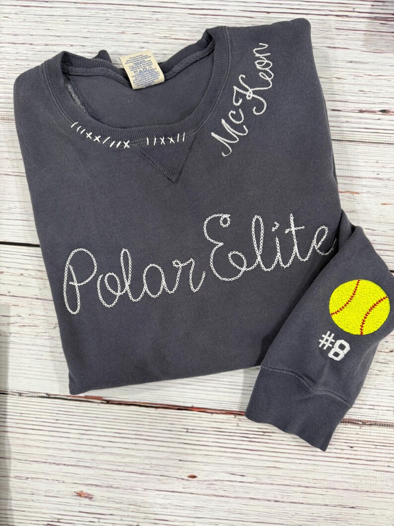 May include: A dark grey sweatshirt with white embroidered text reading "Polar Elite" and "McKeon." The sleeve features a yellow softball with red stitching and the number 8. The neckline has decorative white stitching.