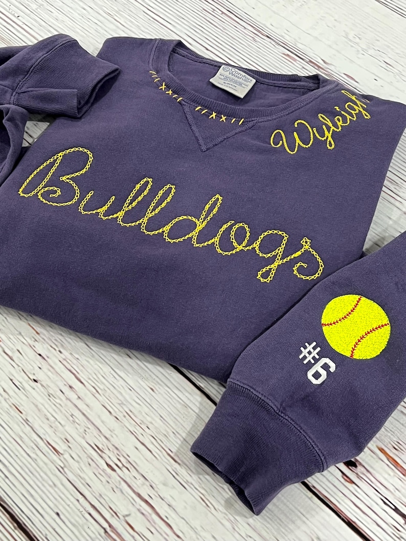 May include: A purple sweatshirt with yellow embroidered text reading "Bulldogs" and "Wyleigh". The sleeve displays a yellow softball graphic and the number "6". The neckline has decorative stitching.