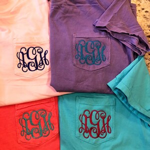 May include: Four t-shirts with a pocket on the front. The shirts are in pink, purple, red, and turquoise. Each shirt has a monogram embroidered on the pocket in a different color.