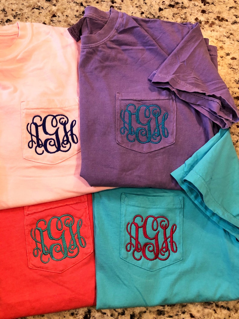 May include: Four t-shirts with a pocket on the front. The shirts are in pink, purple, red, and turquoise. Each shirt has a monogram embroidered on the pocket in a different color.