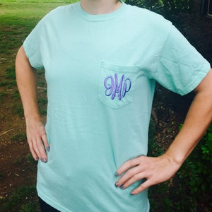 May include: A light blue t-shirt with a pocket on the front. The pocket has a purple embroidered monogram that says "JMP".