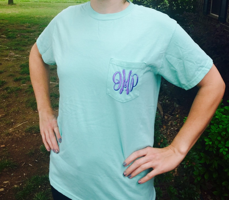 May include: A light blue t-shirt with a pocket on the front. The pocket has a purple embroidered monogram that says "JMP".