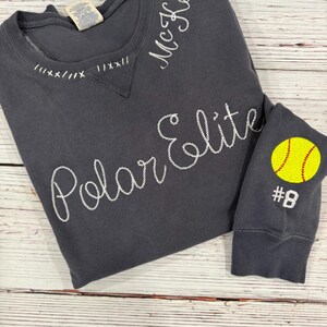 May include: Dark grey sweatshirt with white embroidered text reading "Polar Elite" and "McKeon." The sleeve cuff features a yellow softball with the number 8. The neckline has decorative stitching.