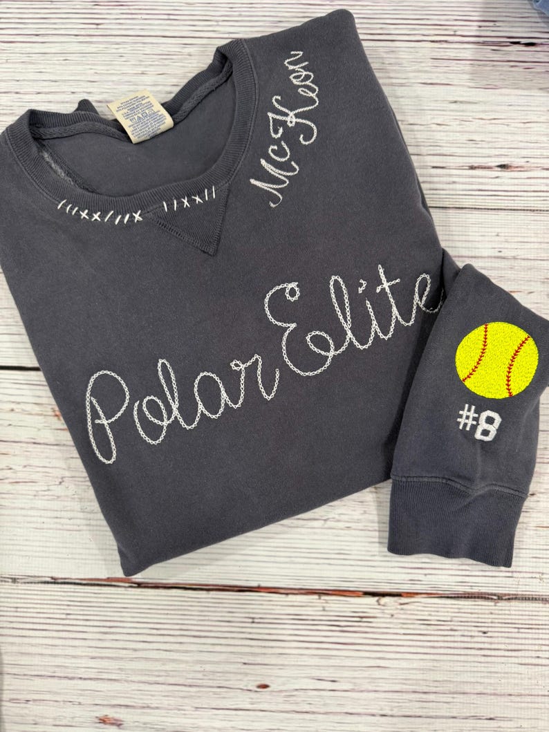 May include: Dark grey sweatshirt with white embroidered text reading "Polar Elite" and "McKeon." The sleeve cuff features a yellow softball with the number 8. The neckline has decorative stitching.