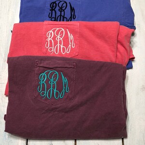May include: Three folded t-shirts with a pocket on the front. The shirts are in different colors: maroon, coral, and blue. Each shirt has a white or teal embroidered monogram on the pocket.