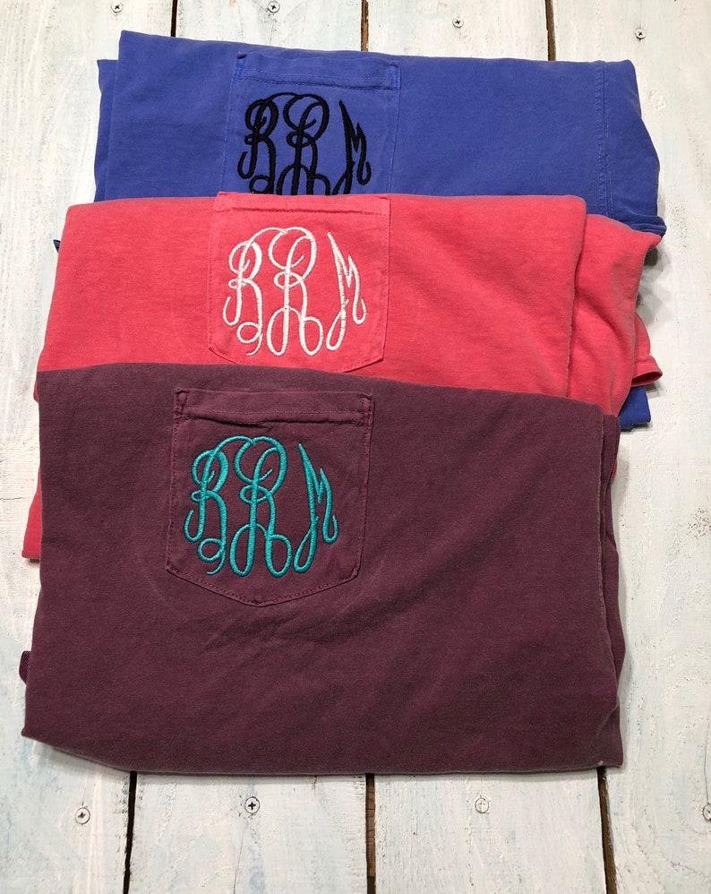 May include: Three folded t-shirts with a pocket on the front. The shirts are in different colors: maroon, coral, and blue. Each shirt has a white or teal embroidered monogram on the pocket.