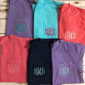 May include: Six t-shirts with different colors and monograms embroidered on the pocket. The colors are purple, light blue, coral, pink, navy blue, and lavender. The monograms are "MSE", "DRR", "HB", "KFG", "MHC", and "SRM".
