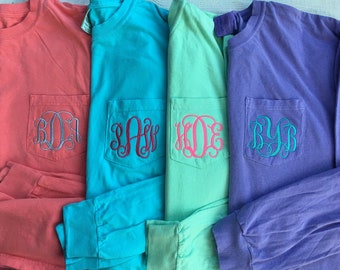 May include: Four long-sleeved shirts in coral, teal, mint green, and purple. Each shirt has a pocket with a different embroidered monogram in a contrasting color. The shirts are neatly folded, showcasing the personalized designs.