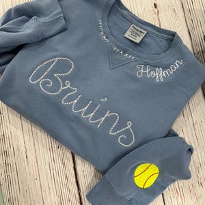 May include: A pale blue sweatshirt with white embroidered text reading "Bruins" and "Hoffman." The neckline has decorative stitching. A yellow softball with red stitching is on the left sleeve. The sweatshirt is made of a soft material.