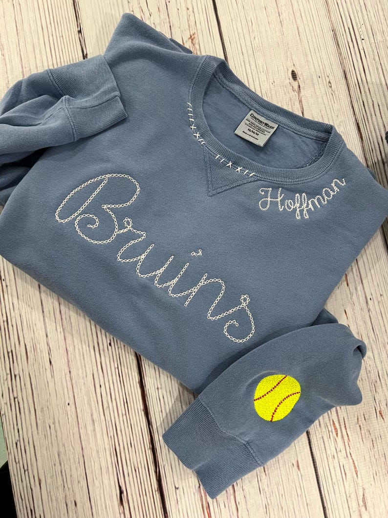 May include: A pale blue sweatshirt with white embroidered text reading "Bruins" and "Hoffman." The neckline has decorative stitching. A yellow softball with red stitching is on the left sleeve. The sweatshirt is made of a soft material.
