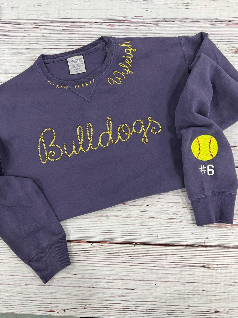 May include: A purple sweatshirt with the word "Bulldogs" embroidered in yellow script across the chest. The name "Wyleigh" is embroidered in yellow on the collar. A yellow tennis ball and the number "6" are embroidered on the left sleeve.