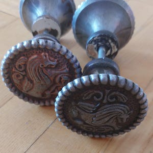 May include: Two antique metal door knobs with a distressed finish. Each knob features a detailed horse design within a decorative border. The knobs have a silver and bronze colour scheme, suggesting an aged appearance. The knobs are likely for interior doors.