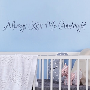 May include: White crib with a white crocheted blanket and a gray mattress. The crib has a white wooden frame and a white metal slatted headboard. The crib is decorated with a stuffed animal and a pillow. The wall behind the crib is light blue and has the text "Always Kiss Me Goodnight" written on it.