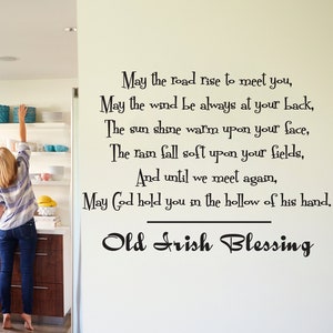 May include: A white kitchen with a yellow door and a woman reaching for a plate on a shelf. The wall has a black and white quote that reads "May the road rise to meet you, May the wind be always at your back, The sun shine warm upon your face, The rain fall soft upon your fields, And until we meet again, May God hold you in the hollow of his hand, Old Irish Blessing."
