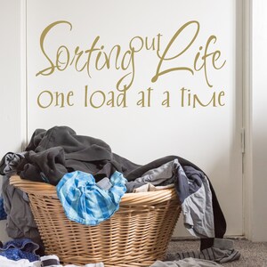 May include: A wicker laundry basket overflowing with a variety of clothing, including a blue plaid shirt, gray pants, and a black sweatshirt. The wall behind the basket has a gold vinyl decal that reads "Sorting out life one load at a time."