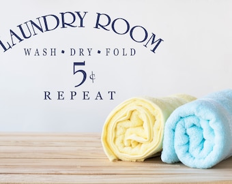 Peut inclure: Un panneau indiquant "Laundry Room Wash Dry Fold 5¢ Repeat" avec deux serviettes roulées, une jaune et une bleue, sur une surface en bois.