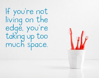 May include: A white ceramic toothbrush holder with five red toothbrushes inside. The text "If you're not living on the edge, you're taking up too much space." is written in blue on a white wall behind the holder.