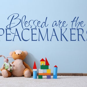 May include: A light blue wall with the text "Blessed are the PEACEMAKERS" in dark blue script. A stuffed teddy bear and a bunny sit on a white rug, with colorful wooden blocks in front of the wall.