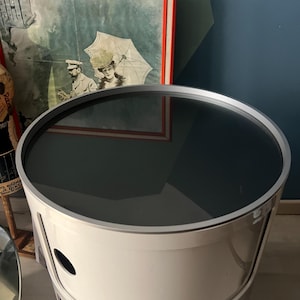 Puede incluir: Una mesa auxiliar cilíndrica blanca con una tapa de cristal oscura y reflectante y un borde plateado. La mesa tiene cortes ovalados y está frente a una impresión vintage enmarcada con un borde rojo. Altura: aprox. 50 cm.