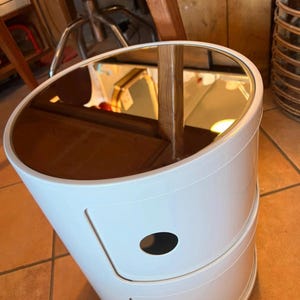 Könnte beinhalten: Weißer, zylindrischer Nachttisch mit Spiegeloberfläche. Der Tisch hat zwei Schubladen mit ovalen Ausschnitten und ein geschwungenes Design. Die Oberseite spiegelt den Raum wider. Der Tisch steht auf einem gefliesten Boden.