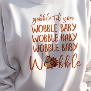May include: Cream-coloured sweatshirt with the phrase "gobble til you wobble baby" in orange script. A cartoon turkey wearing a hat and scarf is featured below the text. The sweatshirt is a casual style.