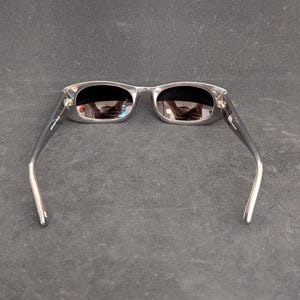 Puede incluir: Gafas de sol con montura plateada y lentes oscuras. Las monturas rectangulares tienen un dise&ntilde;o sutil y moderno. Las patillas son de color gris oscuro y se curvan elegantemente hacia las patillas. Las gafas de sol est&aacute;n sobre un fondo oscuro.