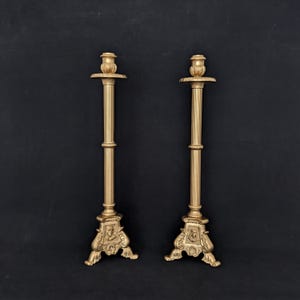 May include: A pair of tall, gold-colored metal candlesticks featuring fluted columns and ornate, decorative bases. Each base is embellished with a small, raised relief portrait of a profile, supported by three scrolled feet for stability.