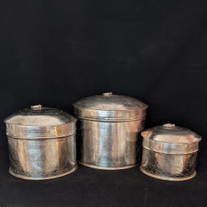 Pode incluir: Três recipientes prateados com tampa de tamanhos variados. Cada recipiente tem um corpo cilíndrico e uma tampa abobadada com uma pequena pega. Os recipientes são decorados com padrões gravados, incluindo formas geométricas e desenhos florais. O fundo é preto.