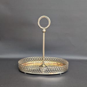 Op de afbeelding: Een zilverkleurig ovaal dienblad met een decoratieve opengewerkte rand en een centrale verticale handgreep met een cirkelvormige ring aan de bovenkant. Het dienblad heeft een gladde, reflecterende bodem en rust op kleine pootjes.