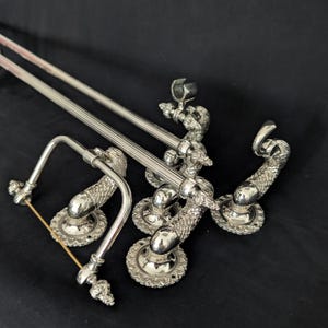 Peut inclure: Ensemble de tringles à rideaux et de quincaillerie décorative en métal argenté. La quincaillerie présente des motifs ornés et détaillés de dragons ou de serpents. Les tringles sont droites et ont une texture striée. Les pièces sont disposées sur un fond sombre.
