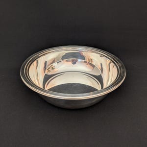 Pode incluir: Uma tigela prateada brilhante com uma borda larga. A tigela tem uma superfície lisa e reflexiva, e a borda tem uma borda sutil e elevada. A tigela é redonda e parece ser feita de metal. A tigela é provavelmente para servir ou exibir.
