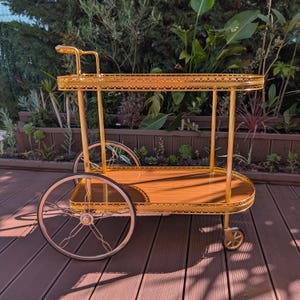 Puede incluir: Un carrito de bar dorado de estilo vintage con dos estantes de madera y ruedas de radios. El carrito tiene un borde decorativo de filigrana y un asa. Diseñado para servir bebidas y entretener.