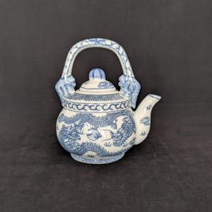 Op de afbeelding: Een witte keramische theepot met blauwe handgeschilderde drakenmotieven die rond het lichaam wikkelen. De theepot heeft een gebogen handvat, een korte tuit en een deksel met een knop. Geometrische patronen sieren de bovenkant van de pot.