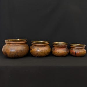 May include: Four antique copper pots with brass rims are arranged in a row, decreasing in size from left to right. The pots have a weathered, aged appearance with variations in color, ranging from dark brown to reddish-brown.