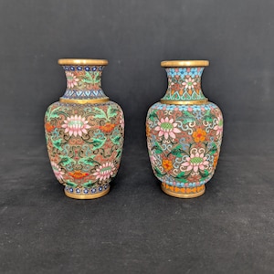Pode incluir: Um par de pequenos vasos cloisonné com um padrão floral detalhado em esmalte rosa, verde, azul e laranja sobre uma base metálica. Cada vaso tem um pescoço estreito, um corpo arredondado e uma borda dourada nas extremidades superior e inferior.